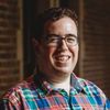 A photo of Garrett, a white man in his mid-20s. He has curly brown hair, glasses, and no beard. He is wearing a brightly colored plaid shirt standing in front of a brick wall